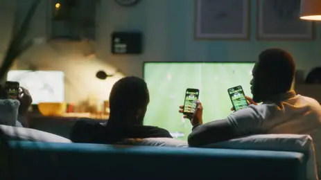 13/07/2026 - Bets - African American man using smartphone and clinking bottles of beer with friends while chilling on sofa and watching soccer game on TV. Foto: Framestock/ Adobe Stock